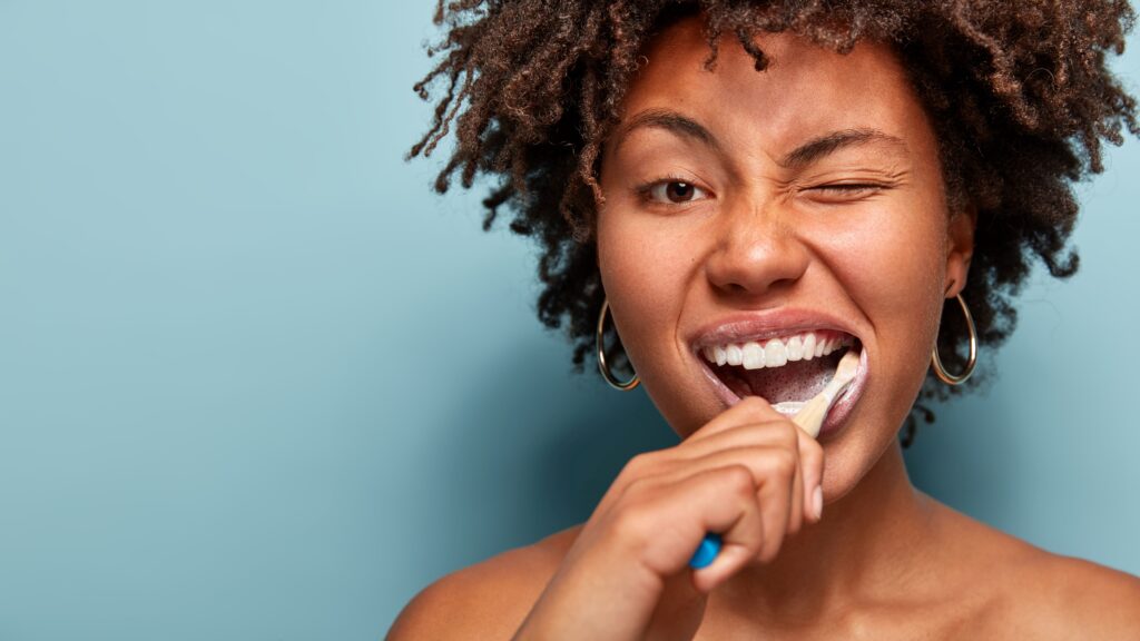 Woman brushing her teeth winking in front of light blue background